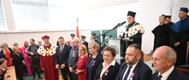 Widok z boku, na podwyższeniu za mównicą stoi mężczyzna w czarnym ceremonialnym stroju z gronostajowym kołnierzem, przed nim w szeregu kilka osób.
