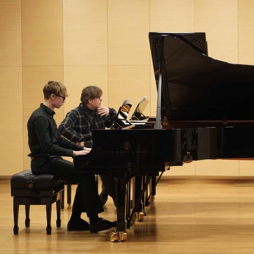 Zdjęcie przedstawia innego ucznia grającego na jednym z fortepianów na scenie sali koncertowej, przy drugim fortepianie prowadzący przysłuchuje się wykonaniu