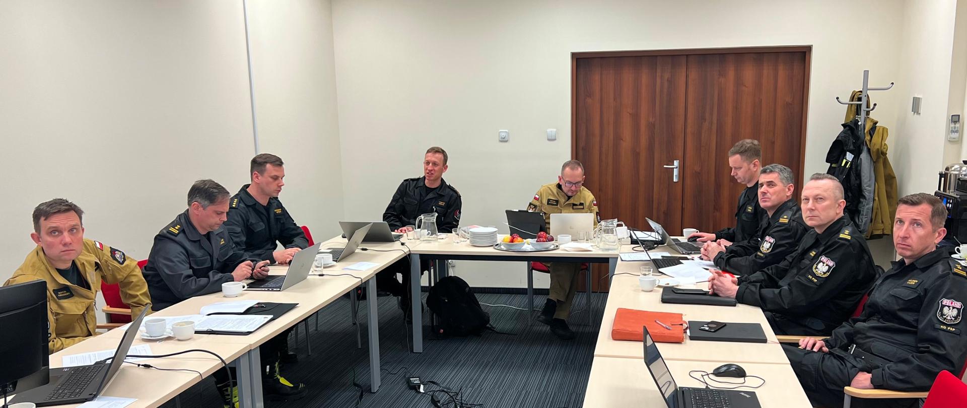 W sali przy stole w kształcie litery U siedzą funkcjonariusze PSP w mundurach koloru piaskowego i czarnego, na stołach znajdują się dokumenty i laptopy 