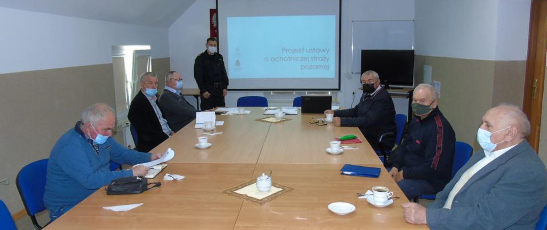W świetlicy komendy powiatowej PSP przy stołach siedzą druhowie z OSP. Na wprost strażak ubrany w czarny mundur dowódczo sztabowy, na ścianie został rozwinięty ekran, na który z rzutnika wyświetlany jest slajd. Wszystkie osoby znajdujące się w pomieszczeniu są w maseczkach. 