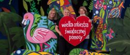  Zdjęcie przedstawia trzy nauczycielki Studium stojące wśród kolorowych obrazków przedstawiających zwierzęta oraz zielonych liści. W rękach trzymają duże czerwone serce z napisem wielka orkiestra świątecznej pomocy.