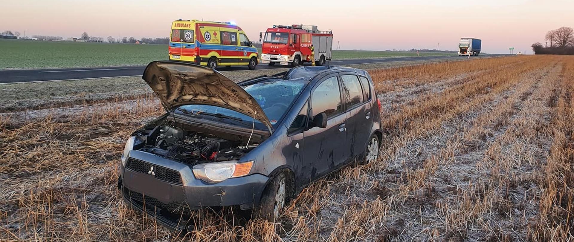 Na zdjęciu widać samochód osobowy po dachowaniu w pozycji na kołach, który stoi na przydrożnym polu. W tle widać samochód pożarniczy oraz karetka pogotowia, które stoją na drodze