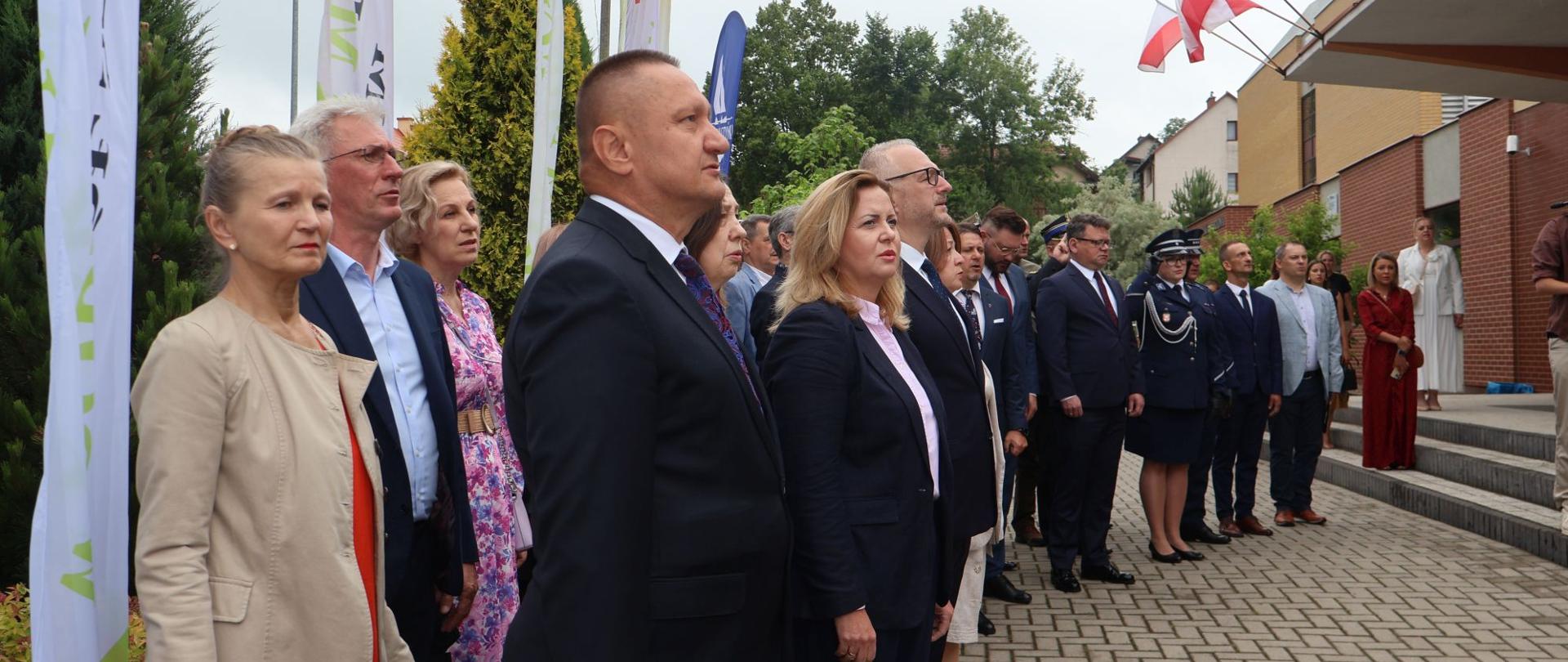 Grupa osób w oficjalnych strojach - mundurach, garniturach, garsonkach stoi na baczność przed budynkiem, na którym powiewają flagi Polski