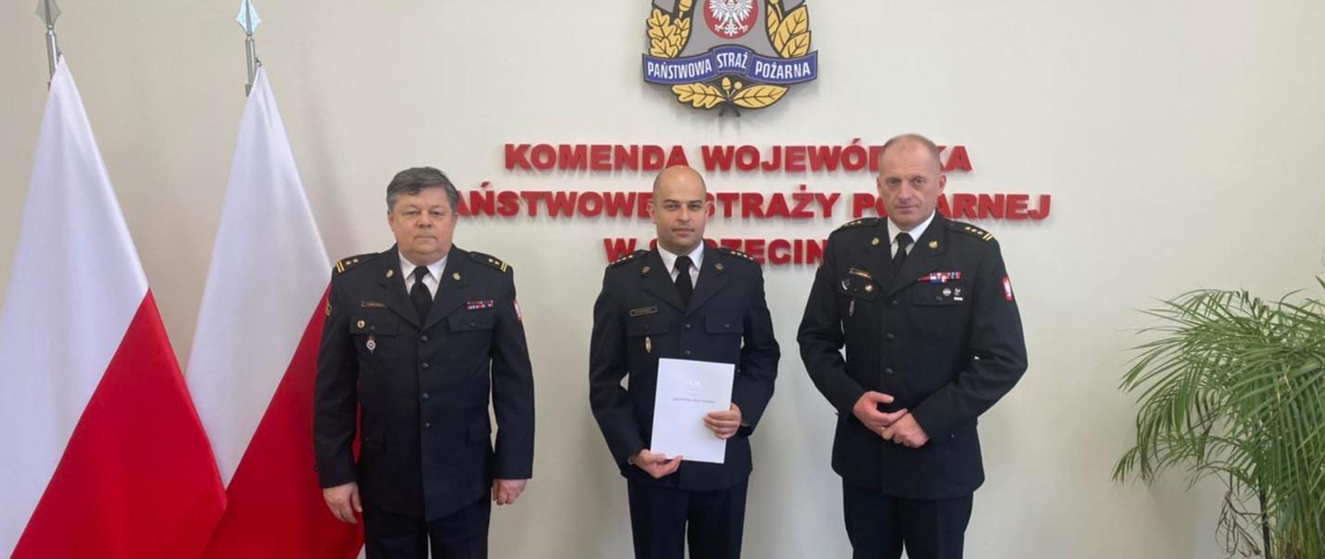 Na zdjęciu trzech funkcjonariuszy, od lewej Komendant Powiatowy PSP w Szczecinku bryg. Andrzej Wójtowicz, Zastępca Komendanta PSP w Szczczecinku st.kpt. Michał Popowicz, Zachodniopomorski Komendant Wojewódzki PSP st. bryg. Jarosław Tomczyk. Po lewej stronie stoją flagi Polski. W tle ściana pamiątkowa KW Szczecin, po prawej stronie kwiat.