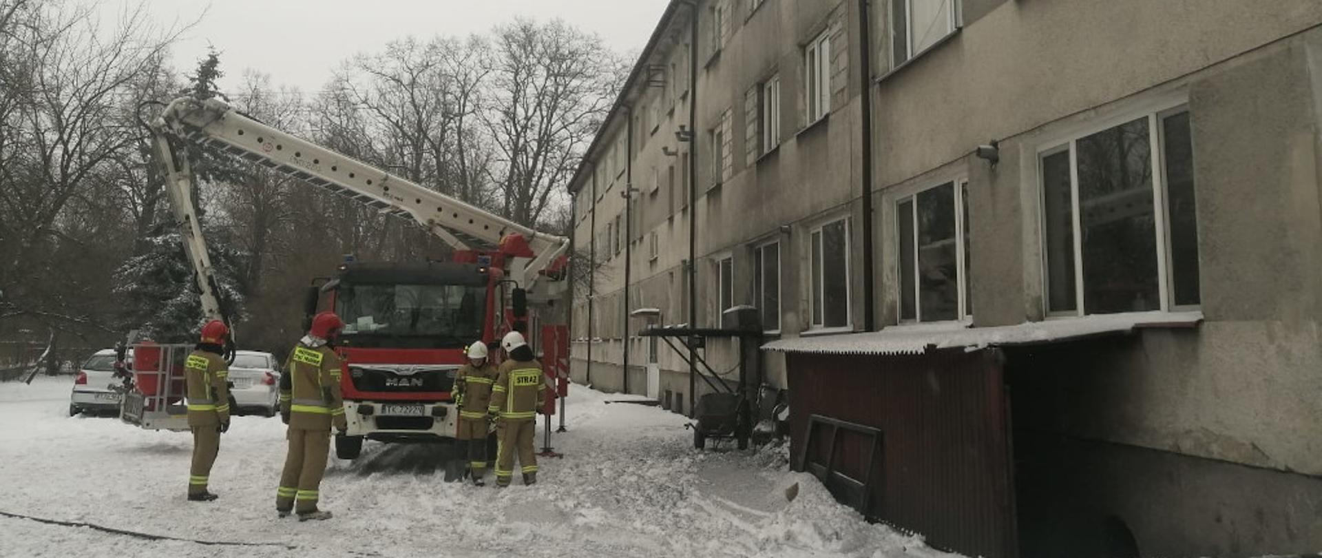 Na zdjęciu przedstawiony jest przód specjalnego samochodu pożarniczego z podnośnikiem hydraulicznym. Pojazd rozłożone ma podpory hydrauliczne oraz przygotowany park drabinowy do wjazdu na dach budynku. Przed samochodem do działań gaśniczych przygotowują się czterej strażacy w umundurowaniu specjalnym. Dwóch z nich na głowie ma założone hełmy koloru czerwonego, natomiast druga dwójka- koloru białego. Po prawej stronie znajduje się frontowa część budynku domu dziecka.