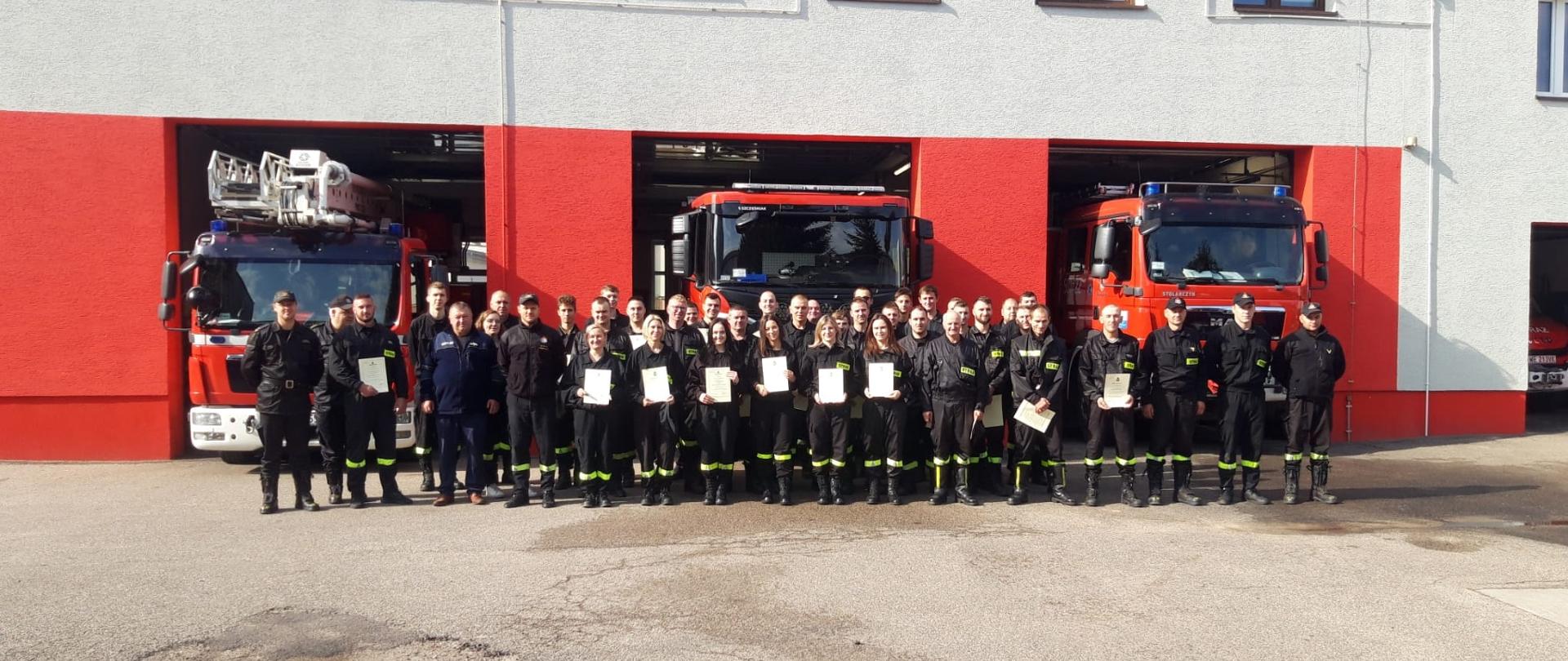 Strażacy OSP stoją z dwuszeregu na placu przed Komendą, w rękach trzymają zaświadczenia. Obok nich stoi komisja egzaminacyjna.