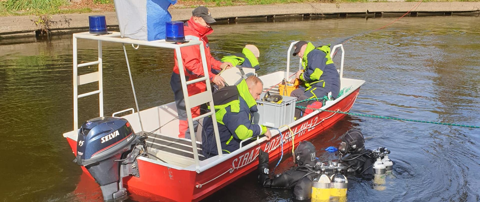 Na rzece czerwona łódź strażacka, w niej strażacy w kombinezonach a w wodzie dwóch nurków.