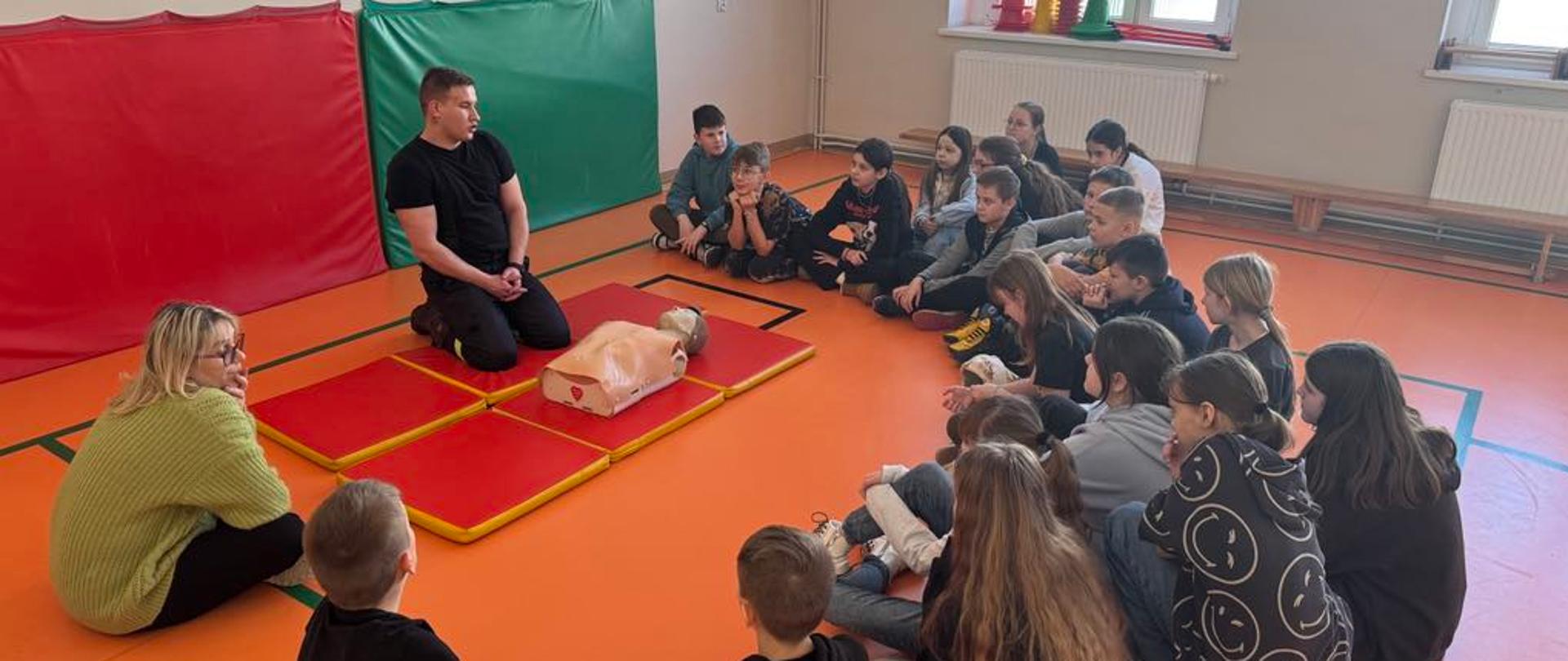 Zdjęcie przedstawia ratownika PSP prowadzącego z grupą dzieci instruktaż pierwszej pomocy