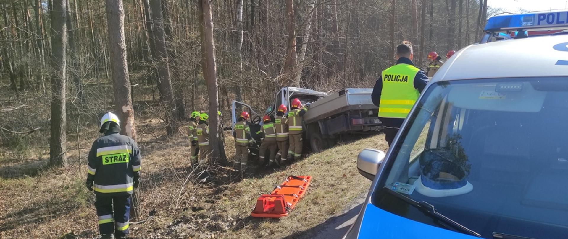Na pierwszym planie radiowóz Policji, nieco dalej Policjant z w kamizelce odblaskowej, w oddali poza drogą samochód który zjeżdżając z drogi uderzył w drzewo, obok samochodu strażacy wykonujący dostęp do kierowcy znajdującego się w pojeździe