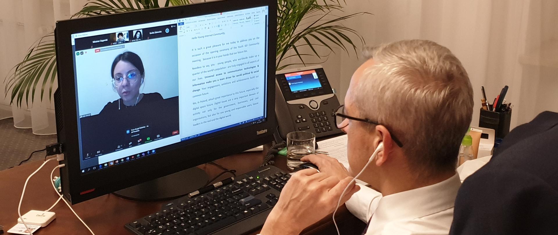 Minister Marek Zagórski siedzi za biurkiem, przed komputerem - w trakcie połączenia i rozmowy z uczestnikami Młodzieżowym Szczytem IGF 2020.