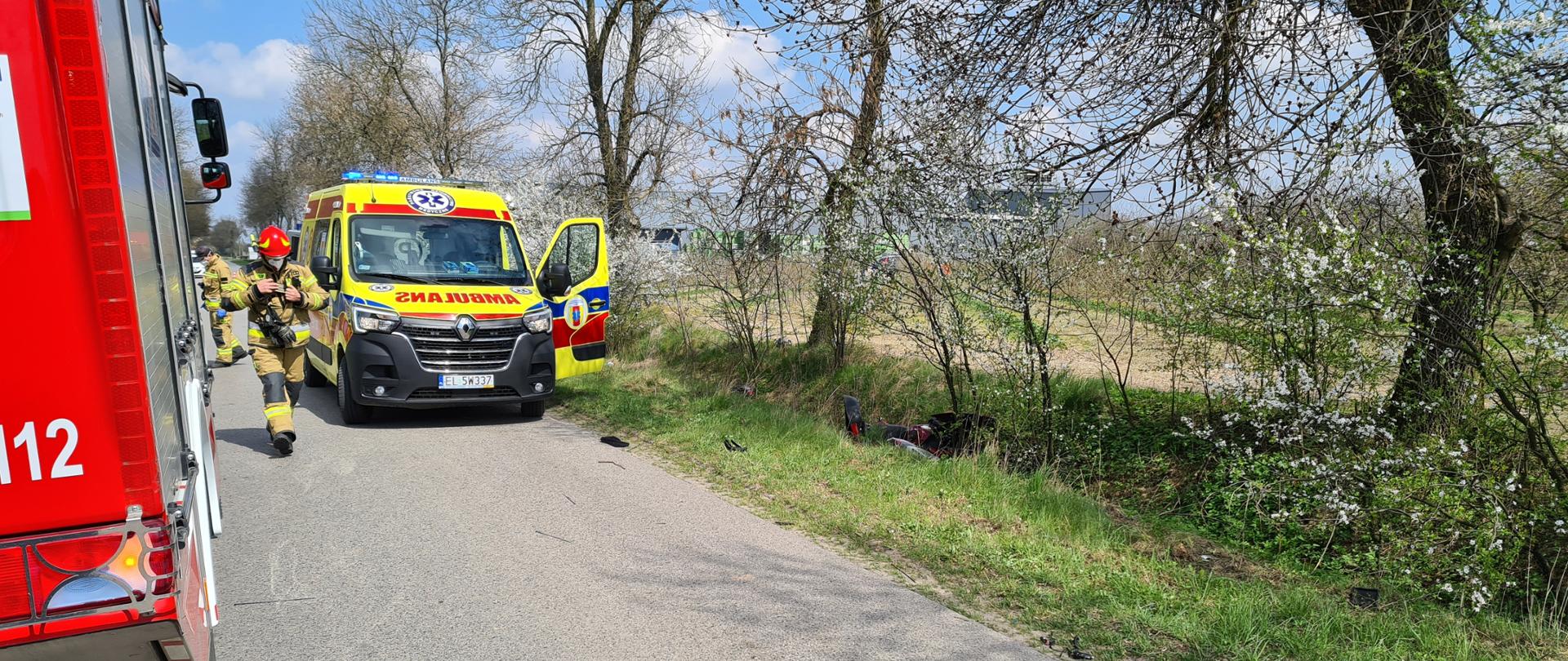 Na zdjęciu widać samochód strażacki oraz samochód karetki pogotowia. Pierwszy z nich stoi tyłem do robiącego zdjęcia, a drugi przodem. Samochody stoją na drodze, na prawo od nich znajduję się rów, w którym widać rozbity motocykl. Między samochodami znajdują się dwaj strażacy.