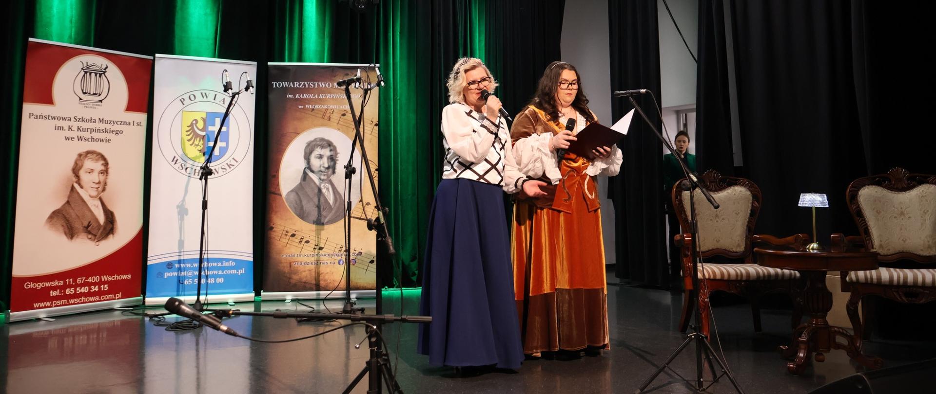 Dwie kobiety w strojach z epoki na scenie. W tle banery PSM we Wschowie, Powiatu Wschowskiego i Towarzystwa Muzycznego.