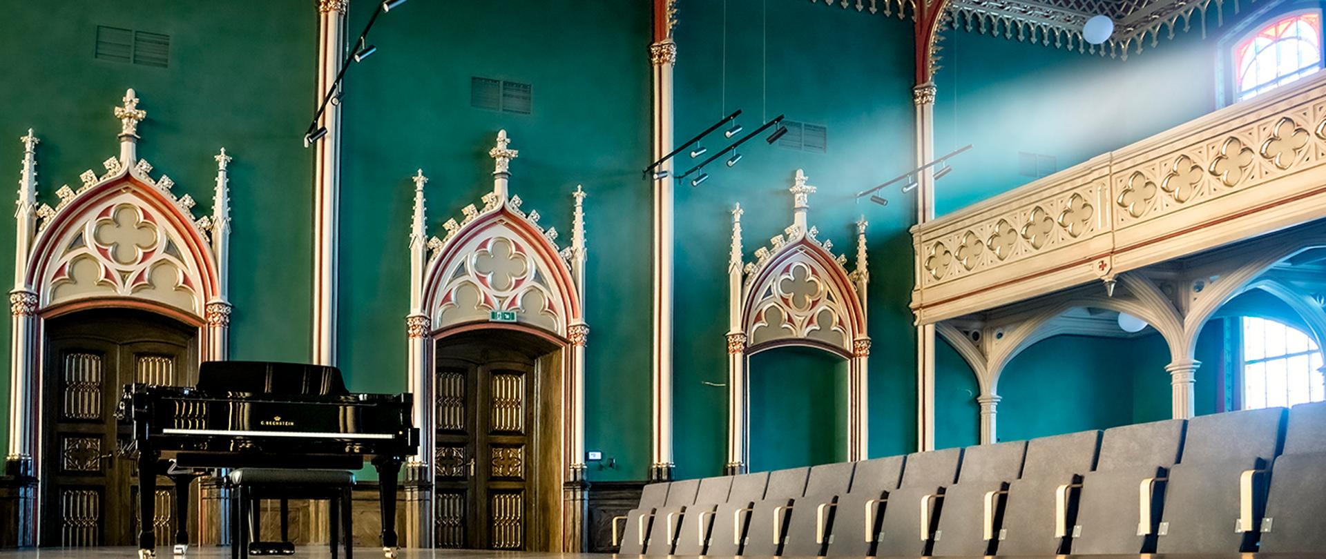 Zdjęcie odnowionej sali koncertowej Ogólnokształcącej Szkoły Muzycznej I i II stopnia im. F. Chopina w Bytomiu. Z lewej strony na pierwszym planie czarny fortepian firmy Bechstein, z prawej rząd jasnych foteli dla publiczności. Na drugim planie widoczna ściana w kolorze ciemno zielonym (butelkowym) z pięknie zdobionymi wnękami w których osadzone są drzwi wejściowe do sali. Drzwi w kolorze brązowym - rustykalnym, bardzo bogato złocone. Z prawej strony widoczny balkon w jasnym kontrastowym do ścian kolorze, ze złoconymi ornamentami przypominającymi czterolistną koniczynę.
Autor fotografii: Arkadiusz Kuc