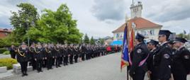 Strażacy w mundurach galowych stoją na szyku na miejscu uroczystości. Po prawej stronie zdjęcia stoją strażacy ze sztandarem. W tle zielone drzewa i duży budynek z czerwonym dachem. 