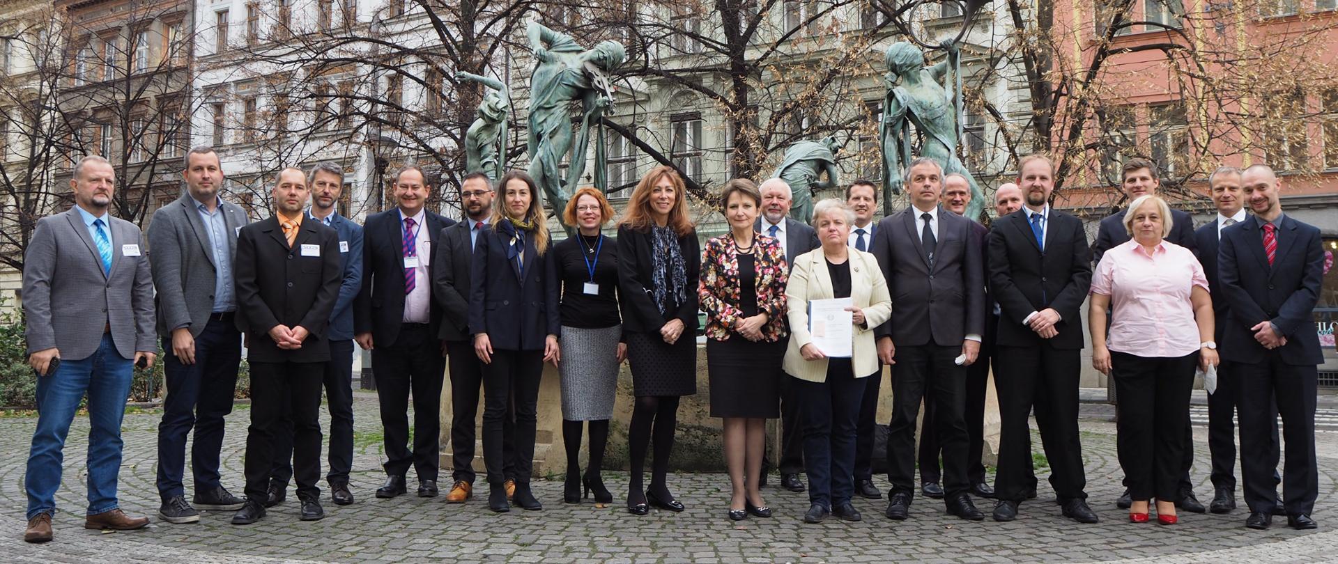 21 osób stoi wybrukowanym placu. Za nimi fontanna z czterema rzeźbami trzymającymi instrumenty muzyczne. W tle jesienne drzewa bez liści i miejskie kamienice. 