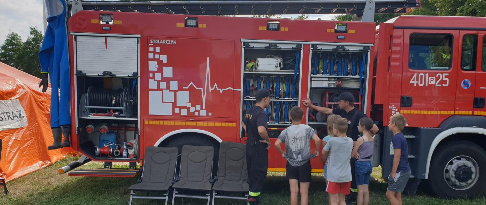 Zdjęcie przedstawia strażaków uczestniczących w festynie profilaktycznym, stojących przy samochodzie gaśniczym i edukujących młodzież szkolną.
W tle samochód gaśniczy oraz drzewa.
