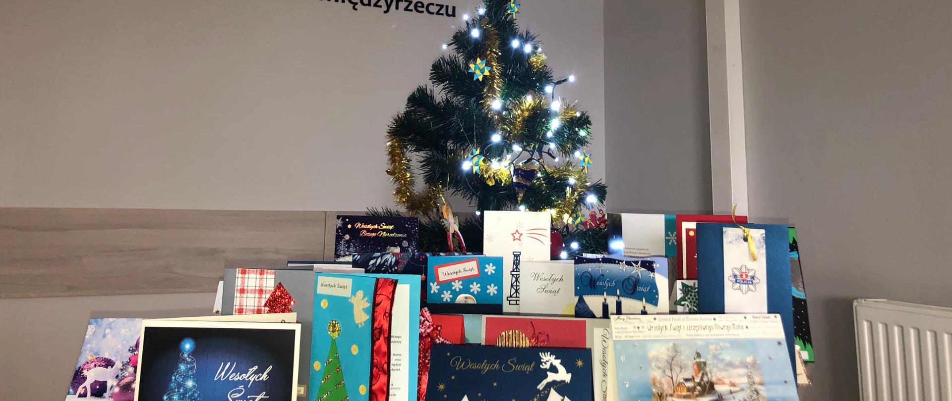 Zdjęcie przedstawia choinkę świąteczną na tle logo Komendy Powiatowej Państwowej Straży Pożarnej w Międzyrzeczu, otoczoną okolicznościowymi kartkami świątecznymi przesłanymi do Komendy Powiatowej z okazji świąt Bożego Narodzenia od firm, instytucji i osób prywatnych.