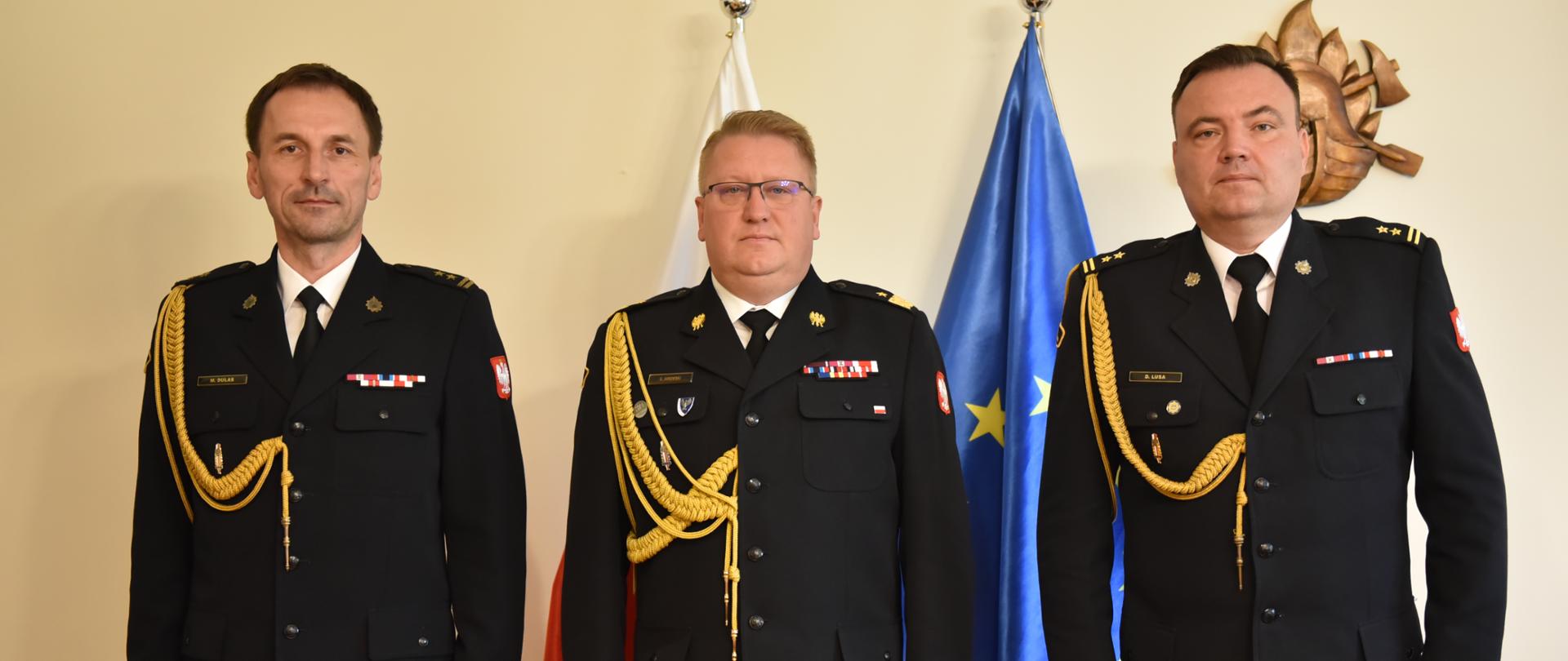 na zdjęciu trzech strażaków w umundurowaniu wyjściowym pozujących do zdjęcia na tle ściany za nimi stoją flaga polski i unii europejskiej 