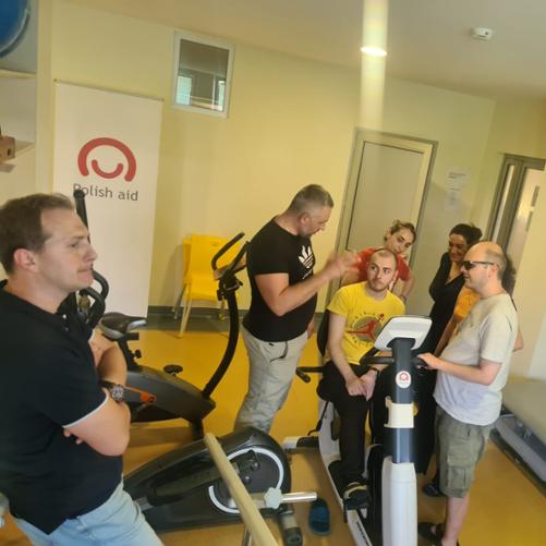 Jedna osoba siedzi na rowerku treningowym z naklejką loga Polska pomoc, 6 innych osób stoi wokół niej i rozmawia, z tyłu widać banner z logo Polish aid