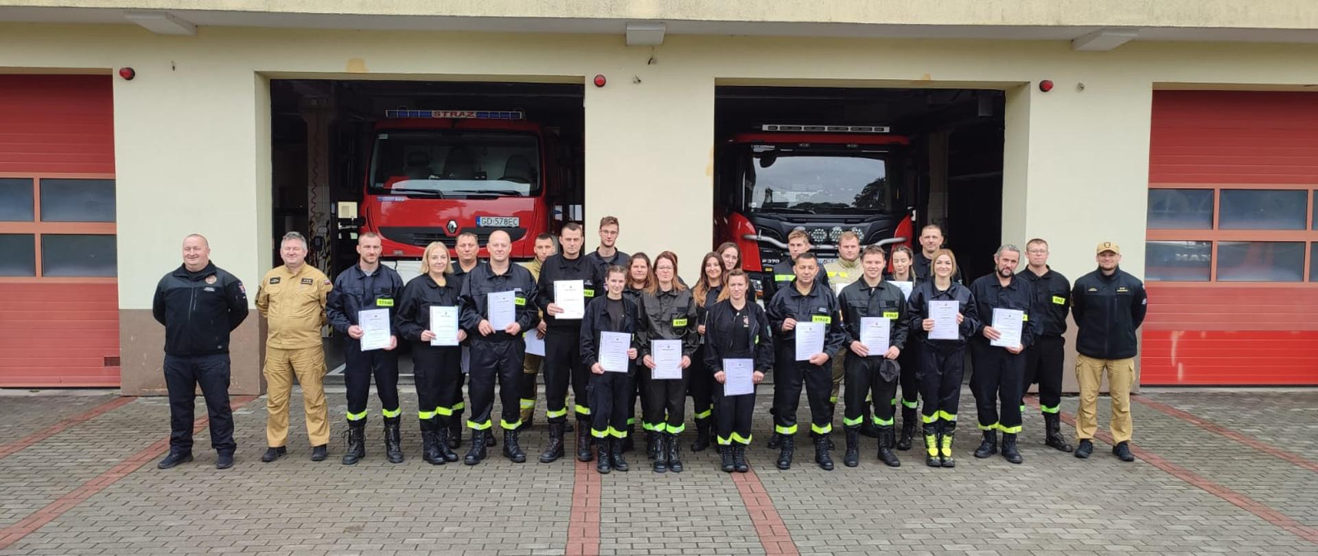 Przed garażami KP PSP w Sztumie stoją ubrani w mundury koszarowe druhowie OSP trzymający w rękach zaświadczenia ukończenia kursu podstawowego. Po prawej i lewej stronie stoi kadra kursu.