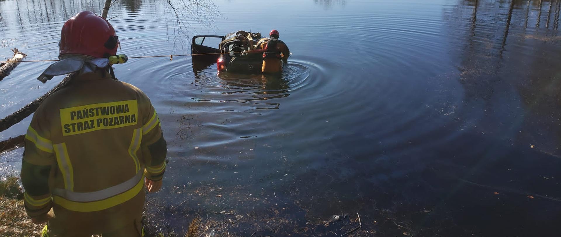 Zdjęcie przedstawia staw, na którego brzegu stoi strażak. W wodzie znajduje się samochód i strażak przygotowujący pojazd do wyciągnięcia na brzeg.