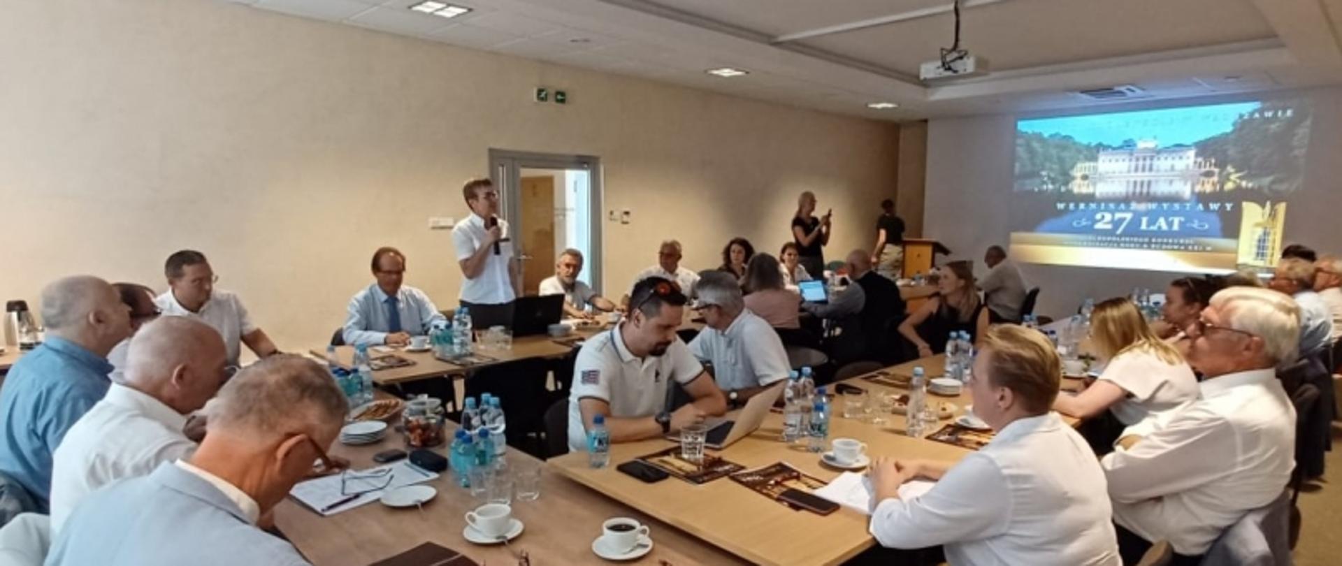 Grupa osób przy stole konferencyjnym, jedna osoba przemawia do mikrofonu; na ścianie ekran z wizerunkiem budynku i napisem „27 LAT”, na stole woda, filiżanki, notatniki i urządzenia elektroniczne.