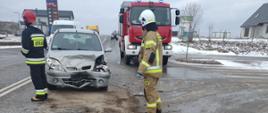Na zdjęciu widać w porze dziennej rozbity przód samochodu osobowego. Przed rozbitym samochodem stoi dwóch strażaków. W tle samochód ratowniczo-gaśniczy. Z auta wyciekają płyny eksploatacyjne. 