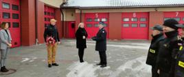Komendant Powiatowy Państwowej Straży Pożarnej w Rypinie wraz z Zastępcą składają podziękowanie i wręczają kwiaty oraz list gratulacyjny księgowej, Pani Joannie Marchlewskiej, za 25 lat wzorowej pracy na rzecz komendy. Odbywa się to na placu jednostki ratowniczo-gaśniczej podczas zmiany służby. Znajdują się tam także pozostali funkcjonariusze Komendy. W tle widać garaże jednostki ratowniczo-gaśniczej.