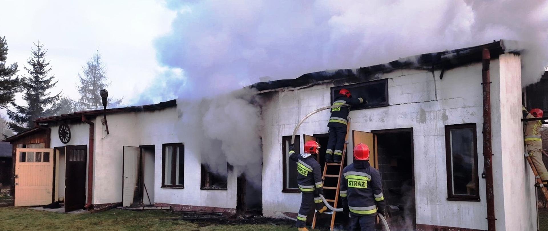 Na zdjęciu widać strażaków na drabinach nasadkowych , podających prąd wody na poddasze palącego się budynku gospodarczego. Są oni zabezpieczani prze kolegów przez podtrzymywanie drabin.