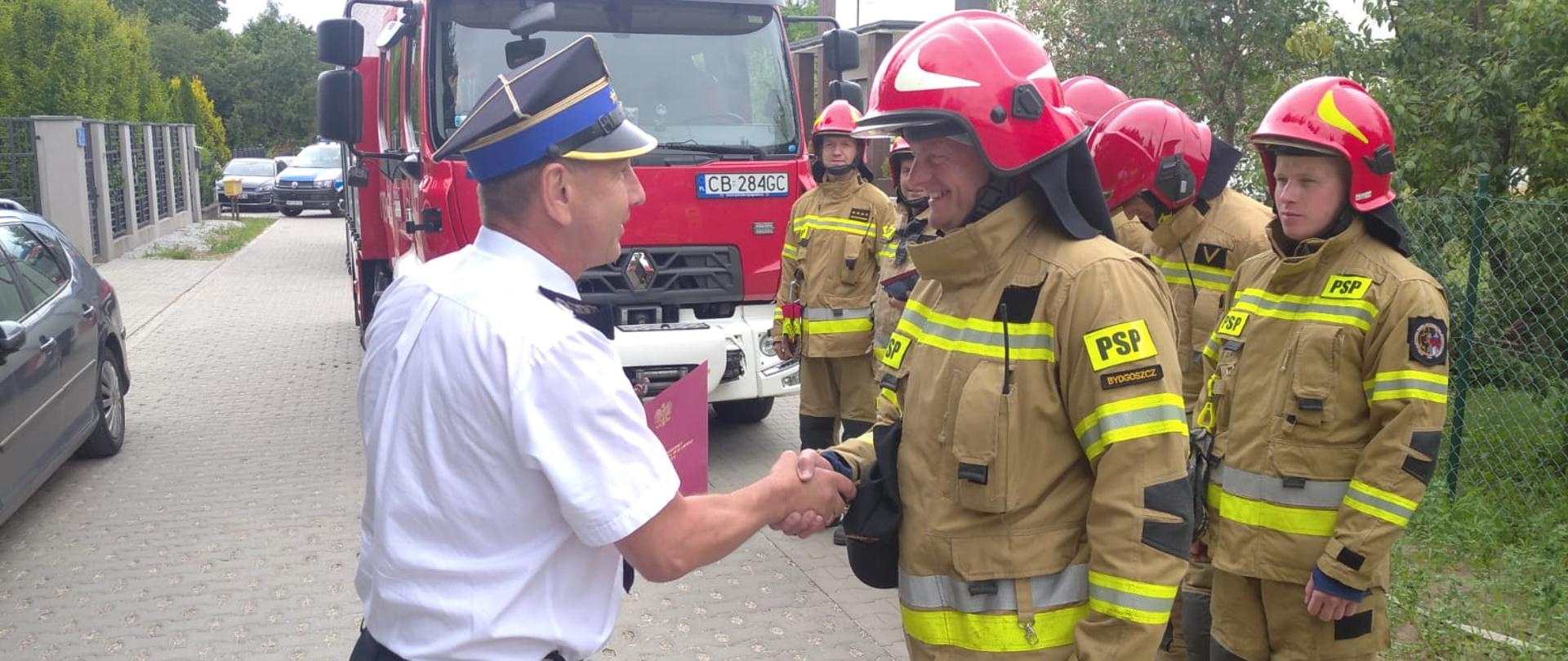 Z lewej Komendant Miejski PSP w Bydgoszczy st. bryg. Janisław Buller w białej koszuli z krótkim rękawem munduru wyjściowego, na głowie czapka rogatywka. Z prawej w żółtym ubraniu specjalnym i czerwonym hełmie na głowie st. ogn. Wojciech Ziemkiewicz. W tle kilku strażaków w żółtych ubraniach specjalnych i hełmach oraz przód samochodu pożarniczego marki Renault. 