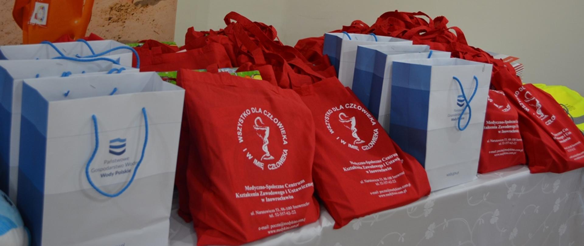 Na zdjęciu torby z nagrodami od Wód Polskich i innych instytucji. Stoją na stole. W tle zdjęcie z bojką ratowniczą. 