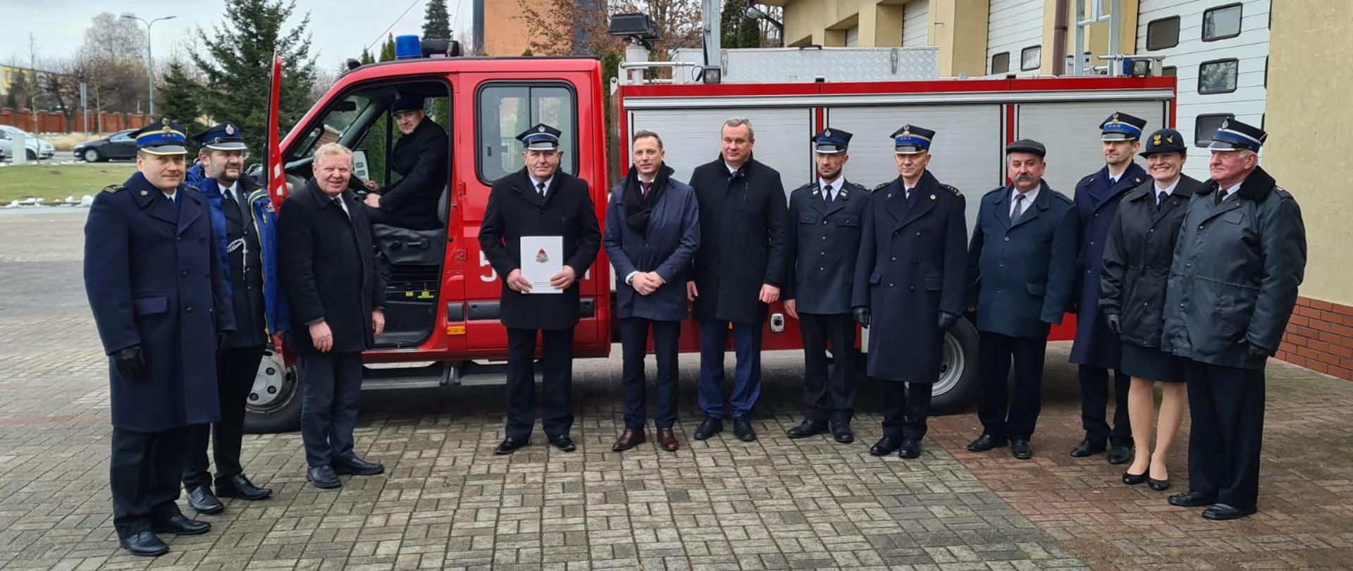 Na placu przed komendą straży pożarnej stoi lekki samochód ratowniczo-gaśniczy Renault Mascott. Obok (na tle) samochodu w jednym rzędzie stoi 12 osób (11 mężczyzn i 1 kobieta) uczestniczących w uroczystym przekazaniu samochodu. 8 osób ubranych jest w mundury wyjściowe, a pozostałe osoby, to cywile, którzy ubrani są w kurtki lub płaszcze zimowe. Jeden mężczyzna siedzi za kierownicą samochodu.