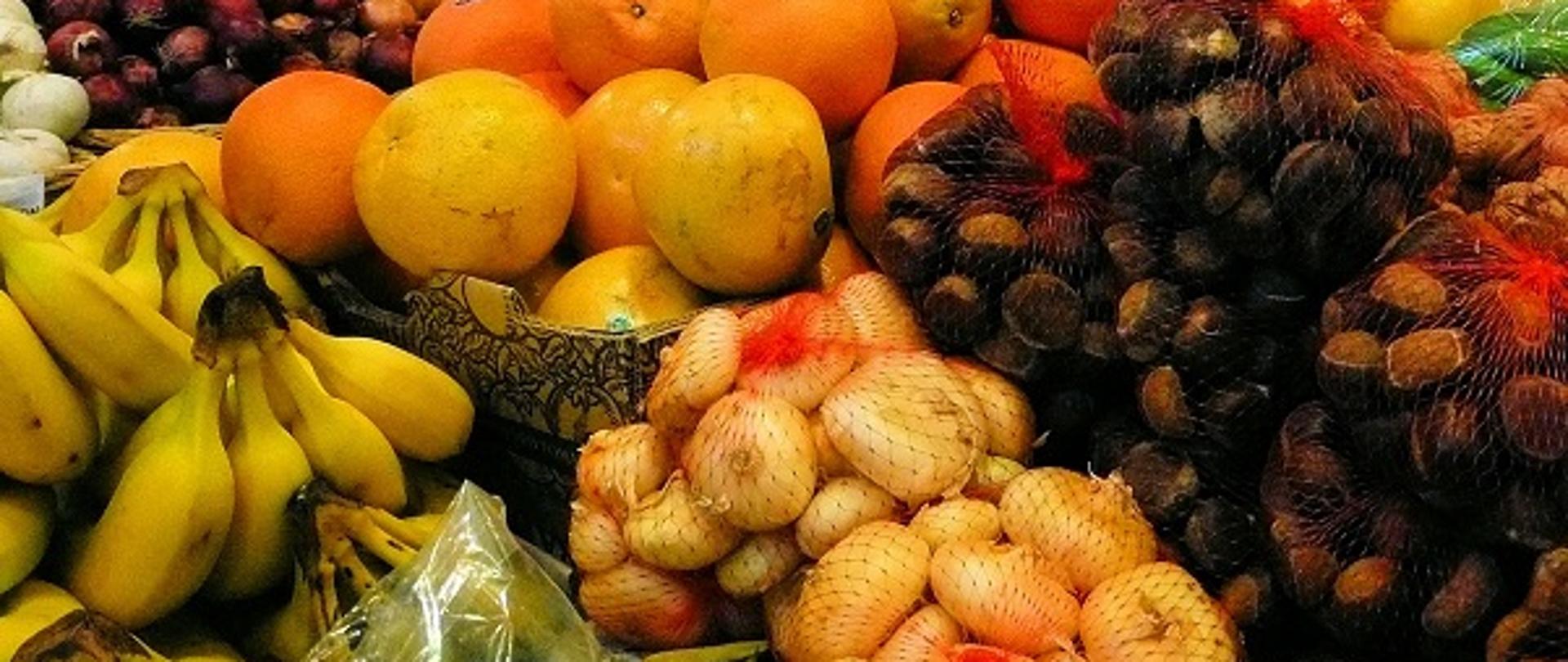 Na zdjęciu są warzywa i owoce leżące na straganie, zapakowane i luzem. Widać cebule, kasztany, pomarańcze, grejpfruty, banany