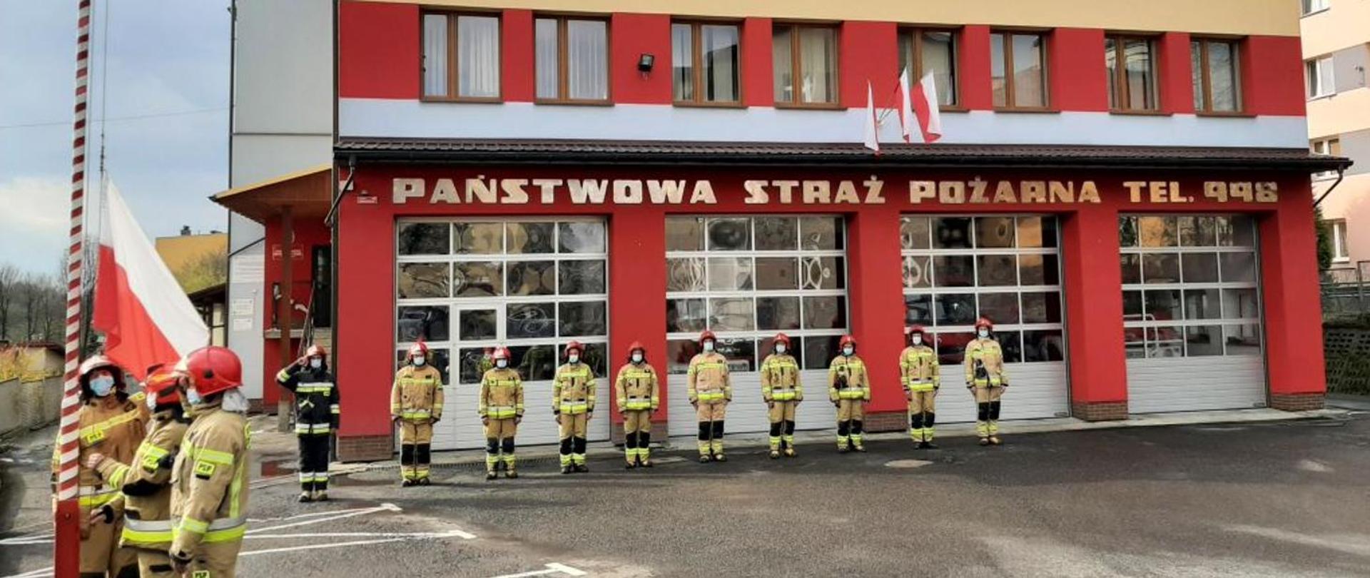 Kolorowe zdjęcie przedstawia uroczystą zbiórkę strażaków w ubraniach bojowych na placu przed budynkiem Państwowej Straży Pożarnej w Brzozowie. Poczet flagowy przy maszcie podnosi biało-czerwoną flagę.
