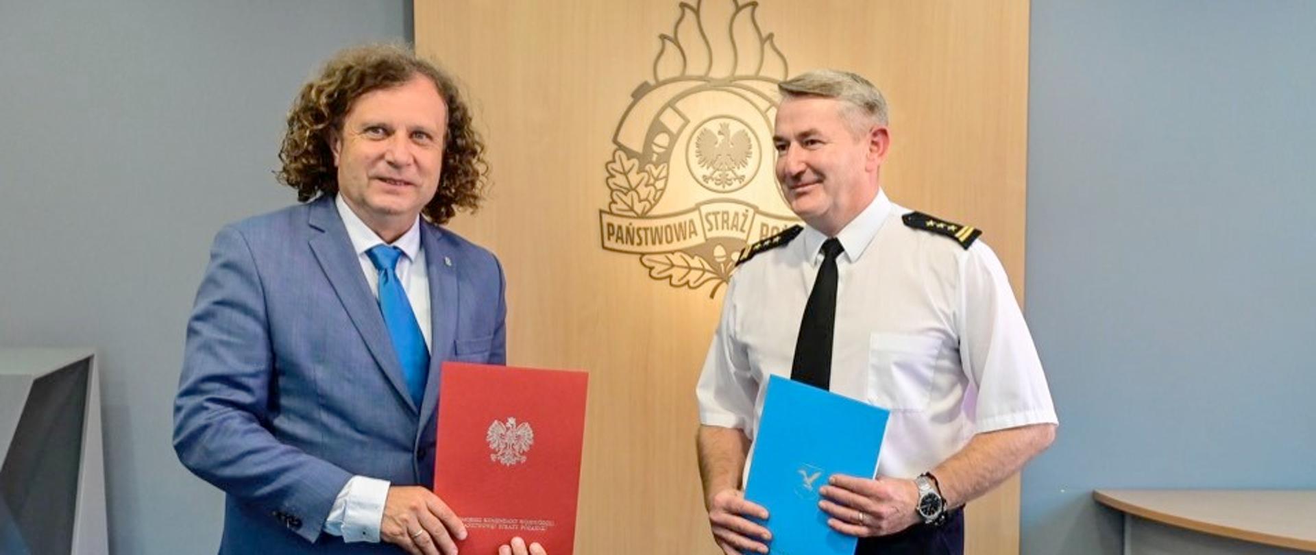 na tle logo PSP przy stole po lewej stronie stoi prezydent sopotu Jacek karnowski trzymając w rękach podpisane porozumienie. Z prawej strony stoi komendant Miejski PSP Sopot st. bryg. Marian Hinca i w rękach trzyma drugi egzemplarz podpisanego porozumienia