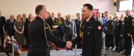 Dwóch strażaków w galowych mundurach podaje sobie ręce podczas uroczystości, w tle zaproszeni goście i funkcjonariusze innych służb
