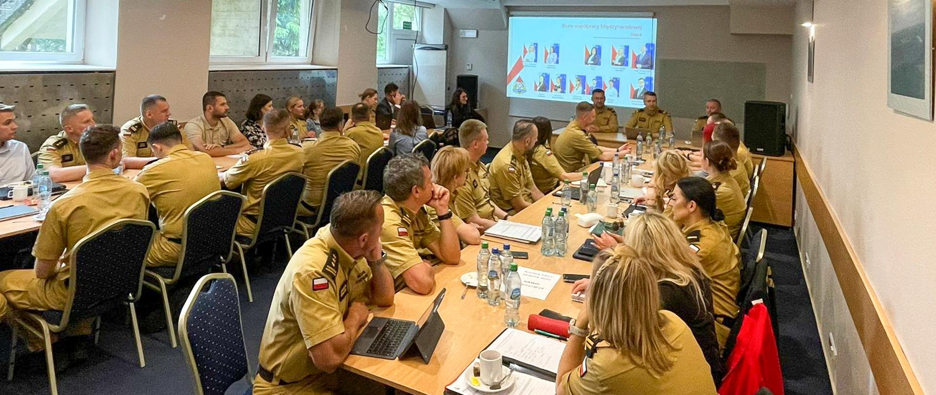 Uczestnicy narady siedzą przy stołach, przed nimi butelki z wodą i laptopy.