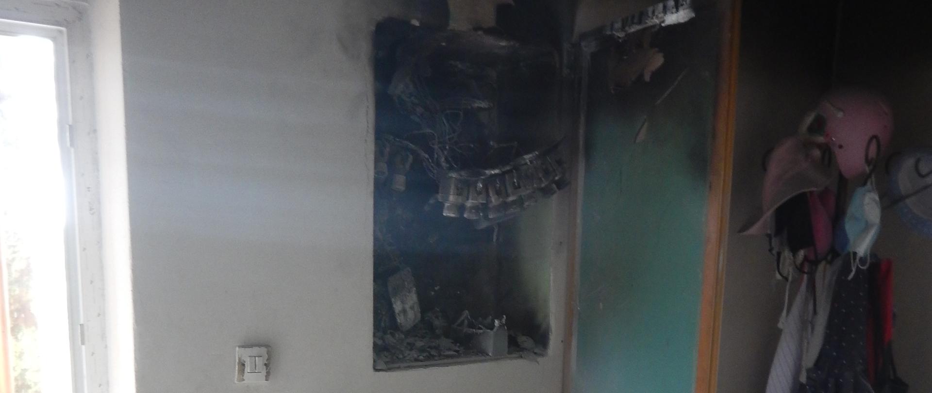 Zdjęcie przedstawia spaloną rozdzielnię elektryczną. W otoczeniu rozdzielni znajduje się gaśnica