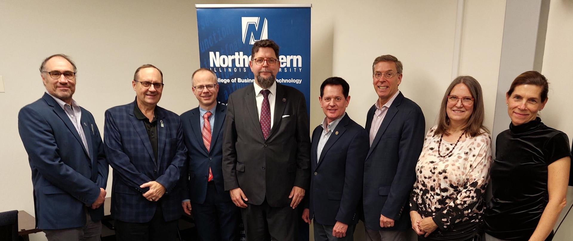 Za szarej podłodze pod granatowym banerem z napisem Northern Illinois University stoi osiem osób, czwarty od lewej wiceminister Szeptycki.