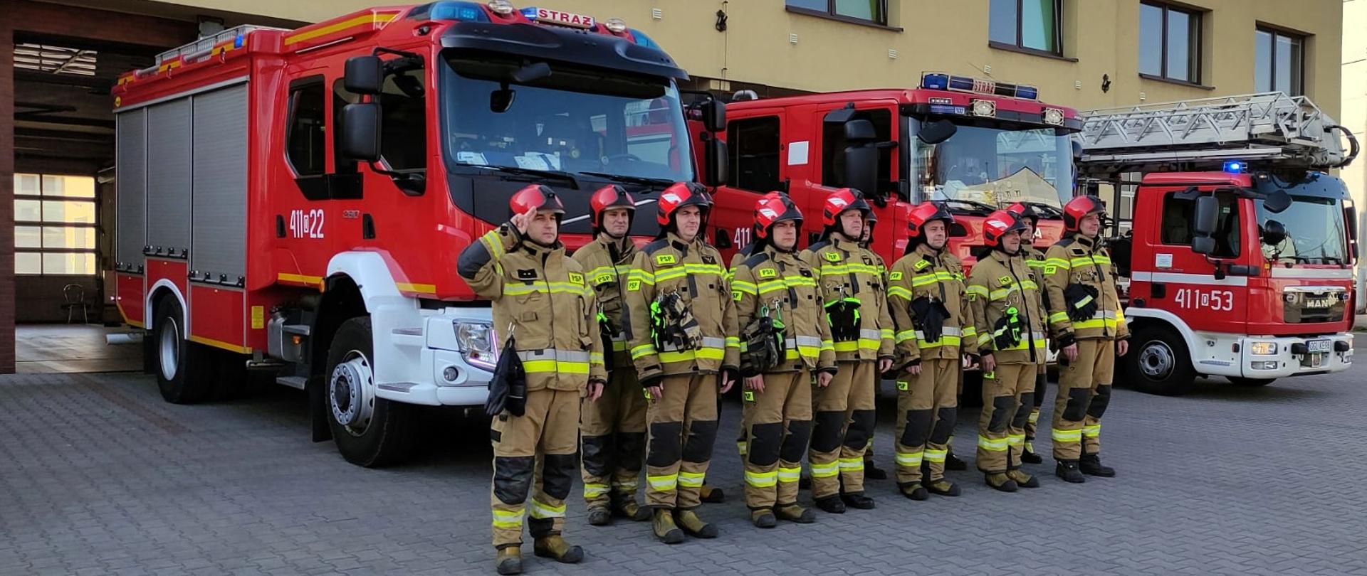 Zdjęcie przedstawia funkcjonariuszy Jednostki Ratowniczo-Gaśniczej Komendy Powiatowej Państwowej Straży Pożarnej w Głubczycach w umundurowaniu specjalnym i hełmach pożarniczych stojących w dwuszeregu przed pojazdami pożarniczymi koloru czerwonego z włączoną sygnalizacją świetlną. W tle widoczny jest budynek strażnicy. Strażacy oddają hołd poległym strażakom w Ukrainie.