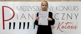 Na zdjęciu znajduje się młoda dziewczynka o jasnych włosach, ubrana w elegancką czarną sukienkę z koronkowymi detalami, białe rajstopy oraz czarne lakierowane buty. Stoi na drewnianej podłodze, trzymając w obu dłoniach dyplom, na którym widnieje napis „DYPLOM” oraz inne szczegóły związane z konkursem pianistycznym. Za dziewczynką znajduje się duży, biały baner z nazwą wydarzenia – „Międzyszkolny Konkurs Pianistyczny Za Kotarą”. Słowo „PIANISTYCZNY” wyróżniono dużymi, stylizowanymi literami, gdzie część z nich imituje klawisze fortepianu. „Za Kotarą” zapisano elegancką, czerwoną czcionką. Baner stoi na czarnym, mobilnym stelażu, a za nim widnieje beżowa, lekko marszczona kotara.