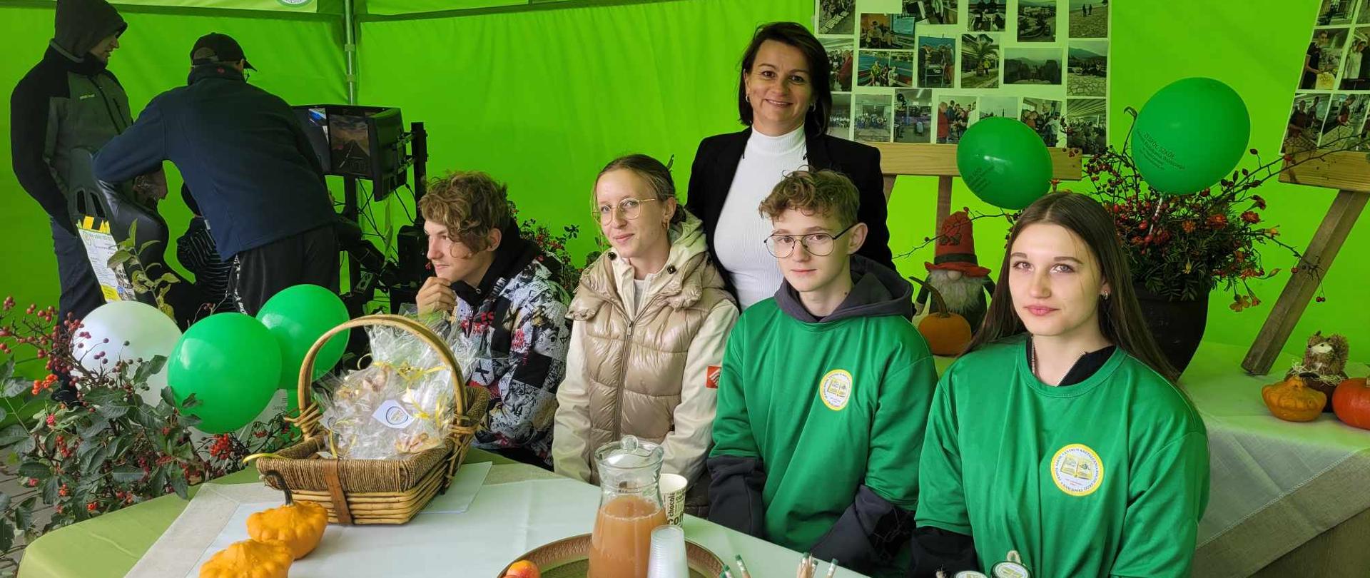 Uczniowie i nauczycielka pozują przy stole w zielonym namiocie. Na stole leżą materiały promocyjne, słoiczki z miodem, szyszki i jesienne dekoracje.