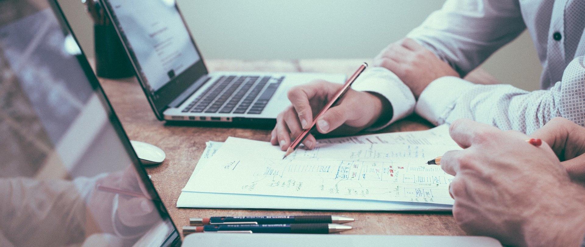 Ilustracja przedstawia laptopy na stole. Przed laptopami widać dłonie osób z nich korzystających.