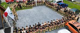 Na zdjęciu widzimy dużą grupę strażaków ustawionych w zwartym szyku w kształcie litery "U". Obok wejścia do budynku powiewają dwie flagi: polska i flaga z logo Lasów Państwowych, co sugeruje, że wydarzenie może być związane z ćwiczeniami przeciwpożarowymi na terenach leśnych.