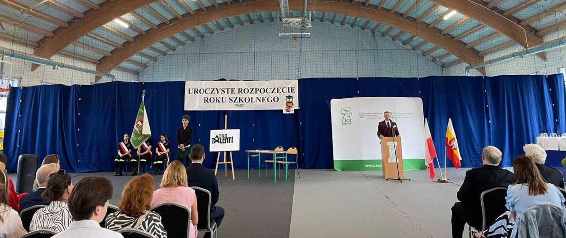 Opis ogólnyZdjęcie przedstawia oficjalną ceremonię rozpoczęcia roku szkolnego, która odbywa się w dużej hali sportowej (prawdopodobnie gimnastycznej). W głębi sali, na tle wielkich, ciemnoniebieskich kotar, zorganizowano scenę. Po prawej stronie za mównicą stoi przemawiający mężczyzna, a po lewej stronie widać poczet sztandarowy. Na pierwszym planie, tyłem do obiektywu, siedzą zgromadzeni widzowie.Architektura wnętrzaSufit: Bardzo wysoki, łukowaty sufit o drewnianej konstrukcji z jasnych belek. Równolegle do łuków biegną długie, jarzeniowe lampy, które jasno oświetlają całą salę. Przez środek sufitu przechodzi gruba, metalowa rura wentylacyjna.Ściana tylna: W tle widać jasnoniebieską ścianę hali, przed którą zawieszono ogromną, pofałdowaną kotarę w kolorze głębokiego granatu, zasłaniającą dolną część pomieszczenia.Scena i dekoracje (środek i głębia kadru)Transparent: W centralnym punkcie, wysoko na granatowej kurtynie, wisi duży, biały baner z czarnym napisem: „UROCZYSTE ROZPOCZĘCIE ROKU SZKOLNEGO”. Poniżej dopisano mniejszym drukiem rok szkolny.Mównica i przemawiający (po prawej): Za drewnianą, jasną amboną (mównicą) stoi elegancko ubrany mężczyzna w garniturze. Przemawia do mikrofonu. Za nim stoi biała ścianka reklamowa (tzw. roll-up) z zielonym paskiem na dole i logo szkoły (widoczne litery ZS CKR). Obok mównicy stoją dwie flagi na drzewcach: biało-czerwona flaga Polski oraz flaga żółto-czerwona.Poczet sztandarowy (po lewej): Grupa uczniów w ciemnych strojach (wyglądających na mundury strażackie lub szkolne) stoi na baczność. Jeden z nich trzyma duży, zielono-biały sztandar szkoły.Elementy na środku: Pomiędzy pocztem a amboną stoi drewniana sztaluga z białym plakatem (widoczny napis „Arena Talentów”) oraz mały stolik szkolny z dwoma zielonymi krzesłami.Widownia (pierwszy plan)Inni uczestnicy uroczystości siedzą w rzędach na czarnych krzesłach, odwróceni tyłem lub profilem do obiektywu.Wśród publiczności widać dorosłych oraz młodzież. Kobiety po lewej stronie są ubrane w eleganckie, letnie bluzki (m.in. w czarno-białe wzory), a mężczyźni po prawej stronie mają na sobie ciemne garnitury.