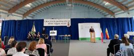 Opis ogólnyZdjęcie przedstawia oficjalną ceremonię rozpoczęcia roku szkolnego, która odbywa się w dużej hali sportowej (prawdopodobnie gimnastycznej). W głębi sali, na tle wielkich, ciemnoniebieskich kotar, zorganizowano scenę. Po prawej stronie za mównicą stoi przemawiający mężczyzna, a po lewej stronie widać poczet sztandarowy. Na pierwszym planie, tyłem do obiektywu, siedzą zgromadzeni widzowie.Architektura wnętrzaSufit: Bardzo wysoki, łukowaty sufit o drewnianej konstrukcji z jasnych belek. Równolegle do łuków biegną długie, jarzeniowe lampy, które jasno oświetlają całą salę. Przez środek sufitu przechodzi gruba, metalowa rura wentylacyjna.Ściana tylna: W tle widać jasnoniebieską ścianę hali, przed którą zawieszono ogromną, pofałdowaną kotarę w kolorze głębokiego granatu, zasłaniającą dolną część pomieszczenia.Scena i dekoracje (środek i głębia kadru)Transparent: W centralnym punkcie, wysoko na granatowej kurtynie, wisi duży, biały baner z czarnym napisem: „UROCZYSTE ROZPOCZĘCIE ROKU SZKOLNEGO”. Poniżej dopisano mniejszym drukiem rok szkolny.Mównica i przemawiający (po prawej): Za drewnianą, jasną amboną (mównicą) stoi elegancko ubrany mężczyzna w garniturze. Przemawia do mikrofonu. Za nim stoi biała ścianka reklamowa (tzw. roll-up) z zielonym paskiem na dole i logo szkoły (widoczne litery ZS CKR). Obok mównicy stoją dwie flagi na drzewcach: biało-czerwona flaga Polski oraz flaga żółto-czerwona.Poczet sztandarowy (po lewej): Grupa uczniów w ciemnych strojach (wyglądających na mundury strażackie lub szkolne) stoi na baczność. Jeden z nich trzyma duży, zielono-biały sztandar szkoły.Elementy na środku: Pomiędzy pocztem a amboną stoi drewniana sztaluga z białym plakatem (widoczny napis „Arena Talentów”) oraz mały stolik szkolny z dwoma zielonymi krzesłami.Widownia (pierwszy plan)Inni uczestnicy uroczystości siedzą w rzędach na czarnych krzesłach, odwróceni tyłem lub profilem do obiektywu.Wśród publiczności widać dorosłych oraz młodzież. Kobiety po lewej stronie są ubrane w eleganckie, letnie bluzki (m.in. w czarno-białe wzory), a mężczyźni po prawej stronie mają na sobie ciemne garnitury.
