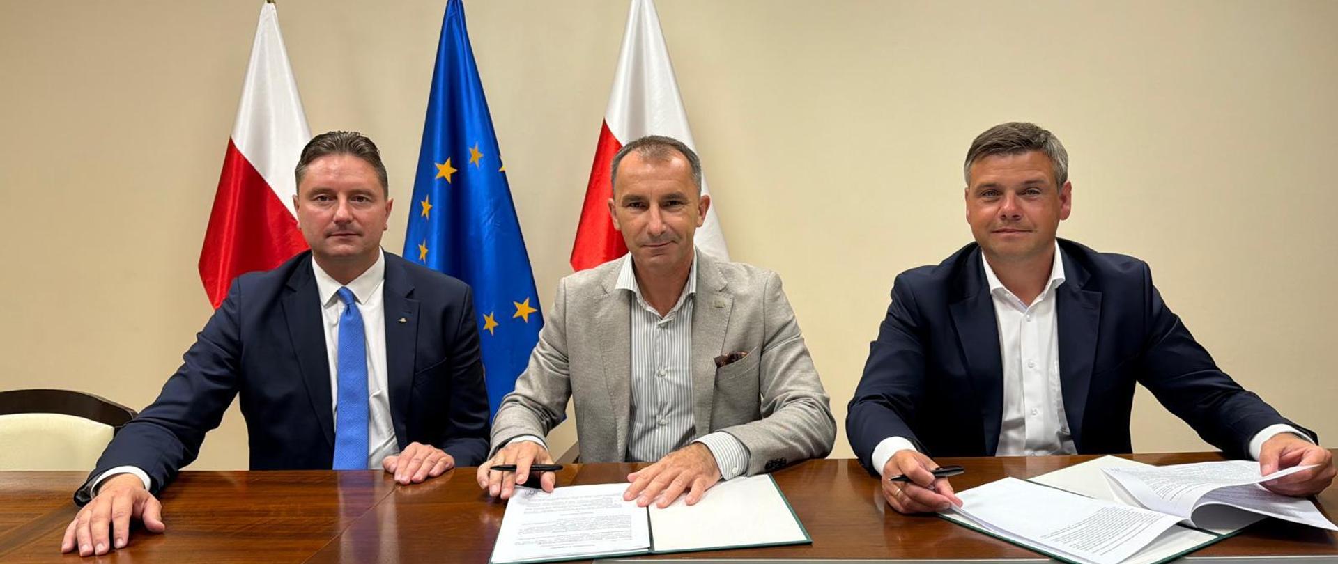 Trzech mężczyzn siedzi przy stole i uśmiecha się do zdjęcia. Dwóch z prawej ma przed sobą ozdobne teczki z dokumentami do podpisu a w dłoniach pióra. Z tyłu za nimi dwie flagi Polski i jedna flaga Unii Europejskiej.