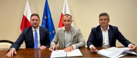 Trzech mężczyzn siedzi przy stole i uśmiecha się do zdjęcia. Dwóch z prawej ma przed sobą ozdobne teczki z dokumentami do podpisu a w dłoniach pióra. Z tyłu za nimi dwie flagi Polski i jedna flaga Unii Europejskiej.