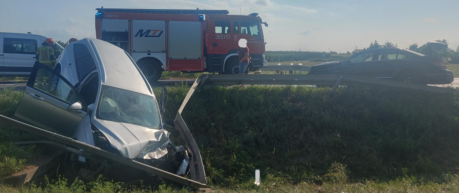 Samochód marki Ford uszkodził barierki ochronne i stoi w rowie z uszkodzonym przodem. Mercedes stoi na jedni i ma otwartą klapę. Z tyłu widać samochód strażacki oraz strażaków. 