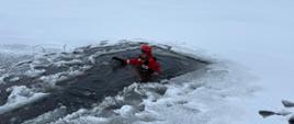 Strażacy w czerwonych ubraniach wypornościowych do pracy w wodzie ćwiczą podejmowanie osoby poszkodowanej, pod którą załamał się lód i wpadła do wody. Na zdjęciu strażacy na saniach lodowych. Zamarznięte jezioro w zimowej aurze. Strażak w wyciętym przeręblu pozoruje osobę poszkodowaną
